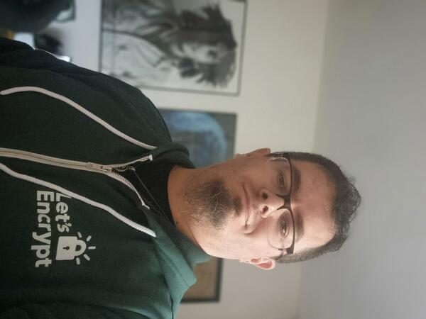 A smiling man with a short hair, glasses, goatee and green hoodie with printed small let's encrypt logo